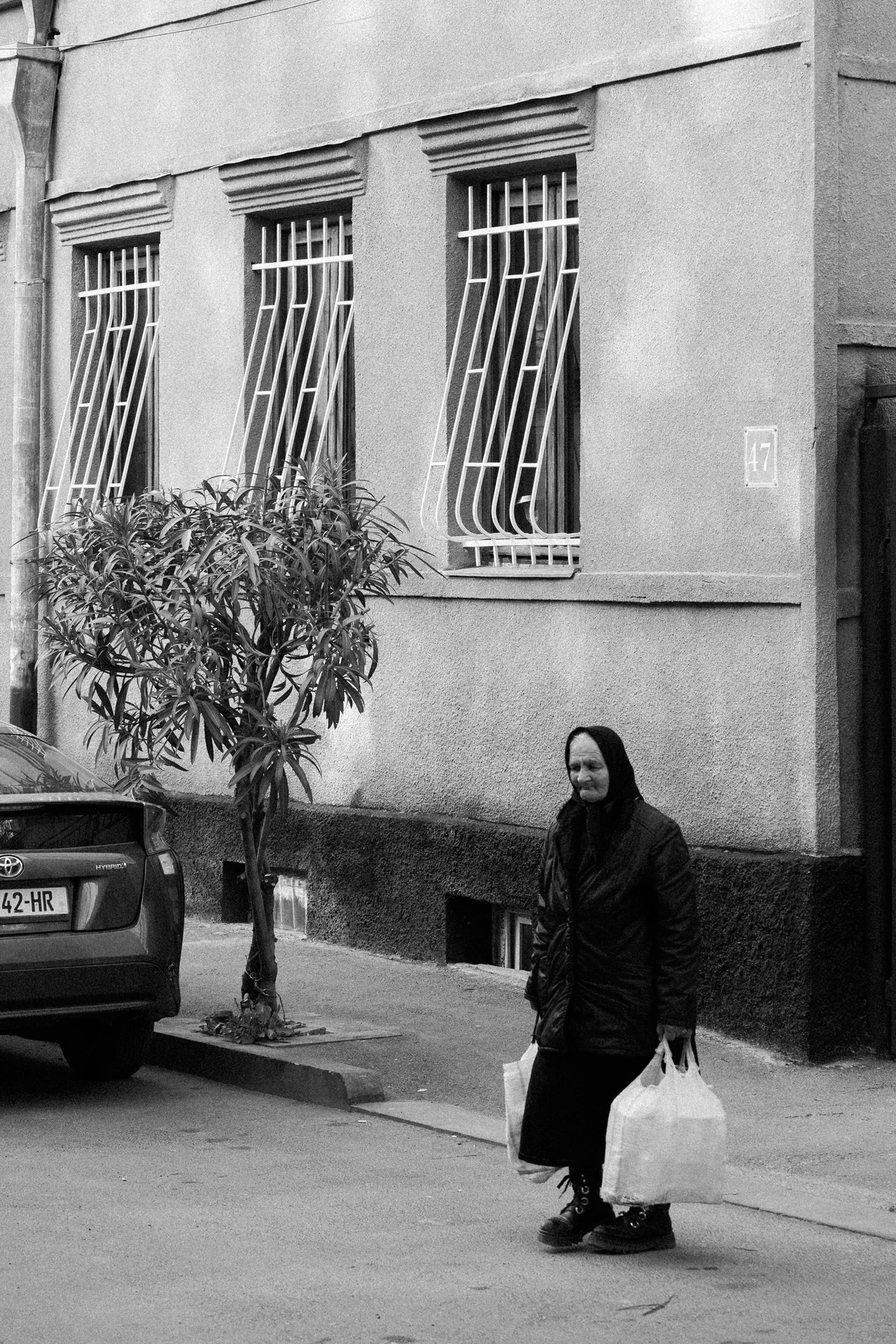Black-and-white street photograph of a woman walking past barred windows