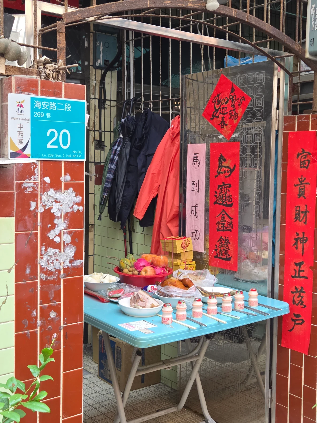 Food offerings on a blue table outside a doorway with red paper decorations and hanging umbrellas