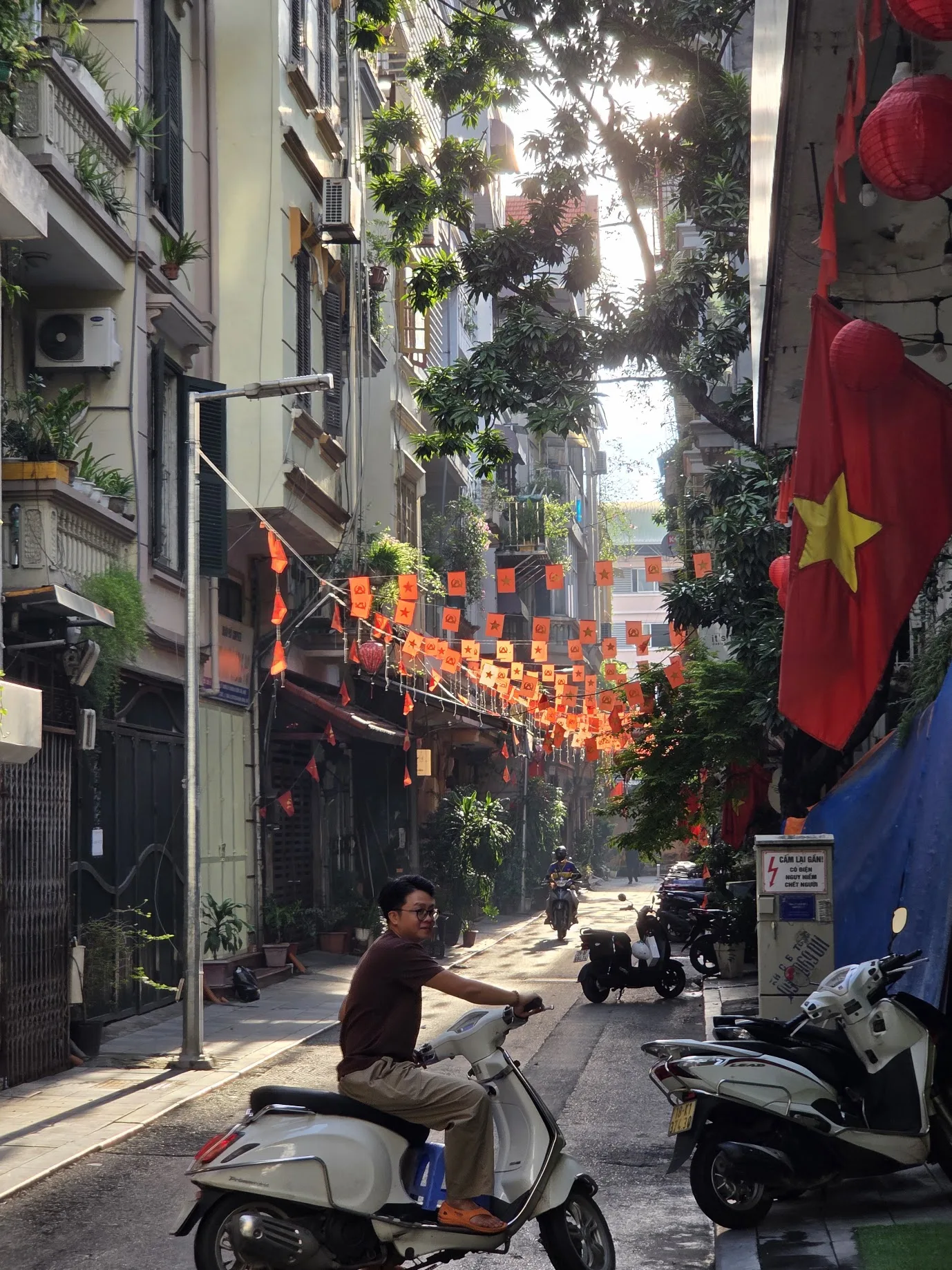 A sunlit Hanoi alley filled with Vietnamese flags and scooters